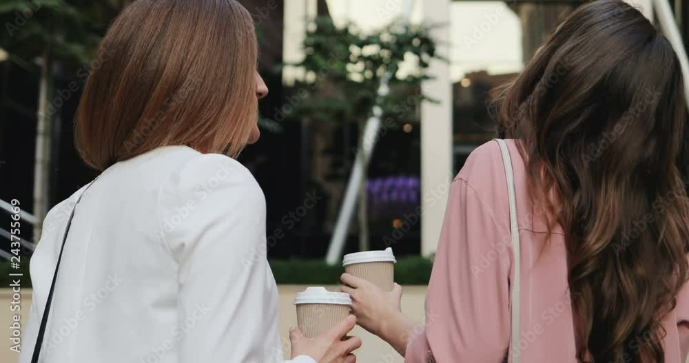 Back view on the two young girls best friends drinking coffee, walking the street and talking cheerfully. Rear. Outdoor. Back view on the two young girls best friends drinking coffee, walking the street and talking cheerfully. Rear. Outdoor.