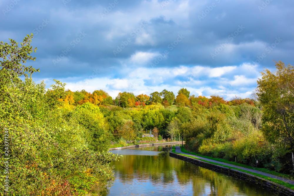 Fototapeta premium Trent and Mersey canal in autumn.Moody sky above woodland and trees with leaves changing colour growing on banks of canal.Idyllic scenery of rural Britain.