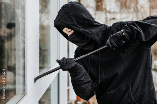 Einbrecher mit Sturmhaube bricht tagsüber in ein Haus durch das Fenster mit einem Brecheisen ein photo