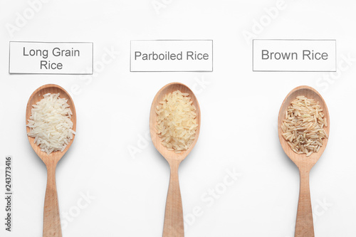 Spoons with different types of uncooked rice and cards on white background, top view