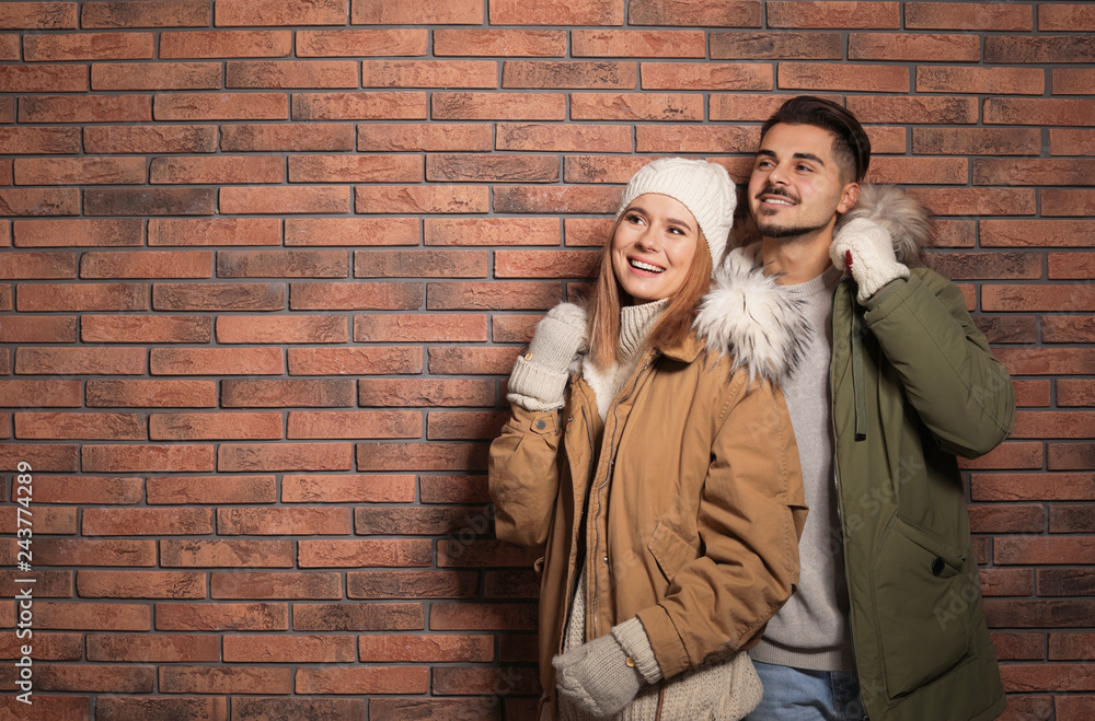 Young couple wearing warm clothes against brick wall, space for text. Ready for winter vacation