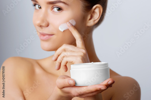 Woman Face Skin Care. Portrait Of Attractive Young Female Applying Cream And Holding Bottle. Closeup Of Smiling Girl With Natural Makeup And Fresh Skin. Beauty Cosmetics.
