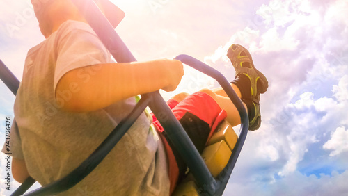 Happy Boy in T-Shirt, Shorts, Baseball Cap and Sandals on Metal Swing with Legs up In the Bright Cloudy Sky on a Summer Day. Low Angle View, Warm Sun Glow