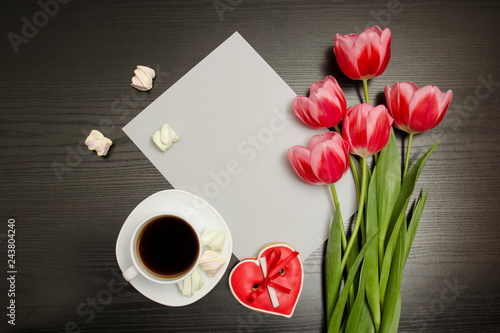 Holiday concept. Bouquet of pink tulips, a cup of coffee, red heart-shaped cookies with a note marshmallow and sheet of paper on a black wooden background. Directly above.