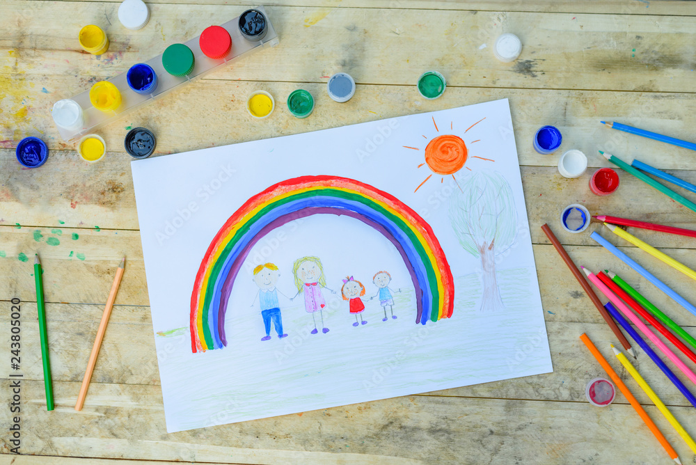 Happy family concept. Drawing on wooden table: father, mother, boy and girl hold hands against background of rainbow and sunny sky.