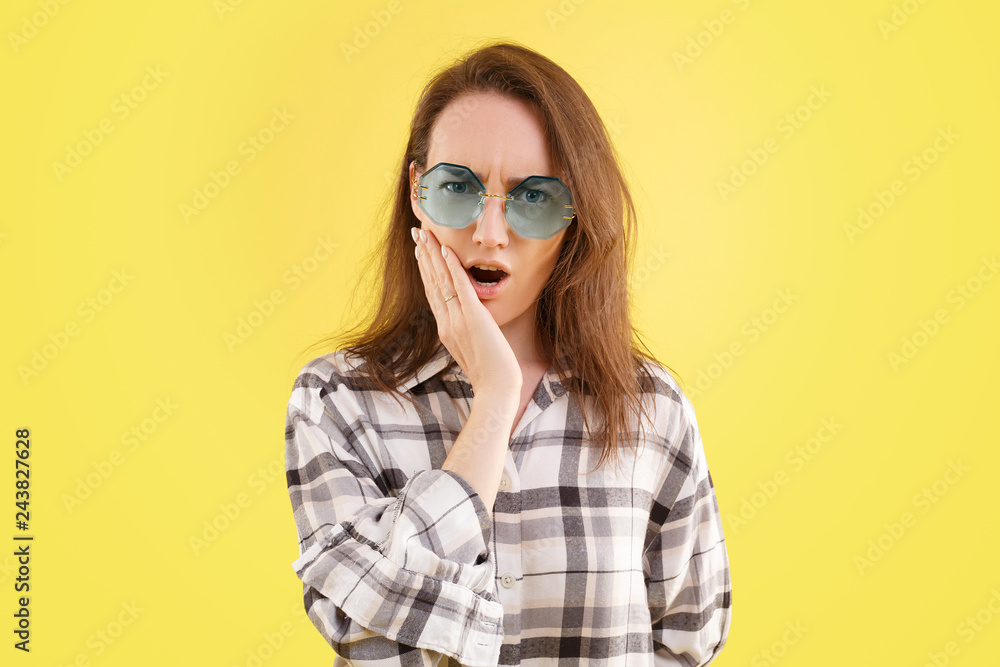 Fototapeta premium Photo of frightened Caucasian woman looks with bugged eyes, keeps mouth widely opened, witnesses terrible accident, has displeased facial expression, in a stylish shirt and glasses, yellow background