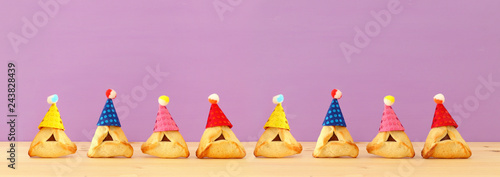 Purim celebration concept (jewish carnival holiday). Traditional hamantaschen cookies with cute clown hats over wooden table and purple background. Banner.