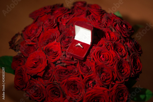 Front view of beautiful wedding ring with precious stone in red velvet gift box on red roses background. For present and marriage proposal.