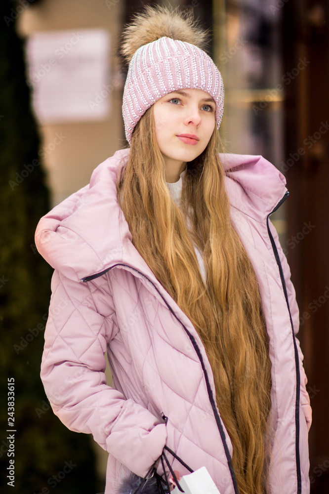 A girl with long blond hair in a pink warm jacket and hat with .pompon stands on the background of golden doors. Winter wonderland. Christmas and New Year concept. A girl with long blond hair in a pink warm jacket and hat with .pompon stands on the background of golden doors. Winter wonderland. Christmas and New Year concept.