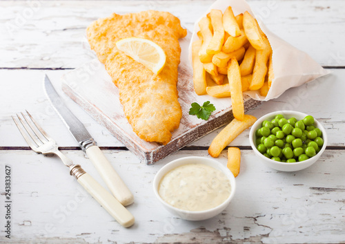 Fototapeta Naklejka Na Ścianę i Meble -  Traditional British Fish and Chips with tartar sauce on chopping board with fork and knife and green peas on white wooden background.