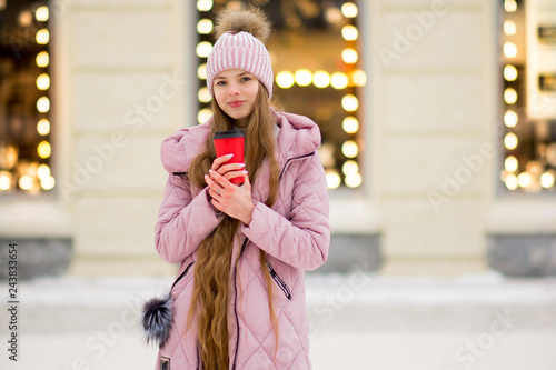 A girl in a pink warm jacket and hat with .pompon stands with coffee in the hand on the background of golden yellow lights and blurred shop windows. Winter wonderland. Christmas and New Year concept.