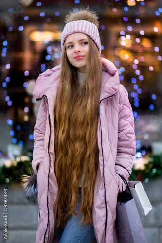 A girl in a pink warm jacket and hat with .pompon stands on the background of multi-colored blue, purple and pink garlands. Winter wonderland. Christmas and New Year concept.