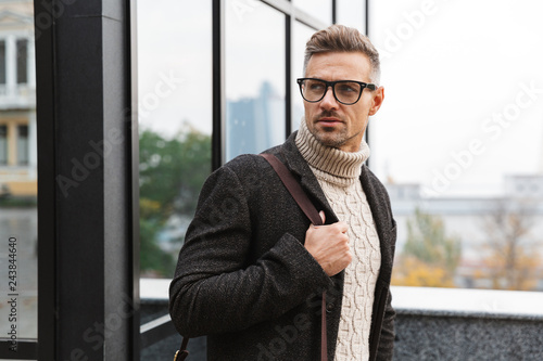 Portrait of stylish man 30s wearing eyeglasses, walking through city street near the building
