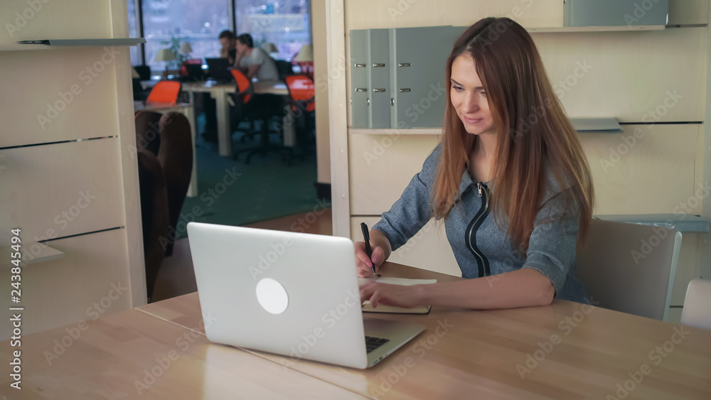 Caucasian model sitting at the light open office. Female writing notes in notebook looking in the laptop. She preparing for meeting. Caucasian model sitting at the light open office. Female writing notes in notebook looking in the laptop. She preparing for meeting.