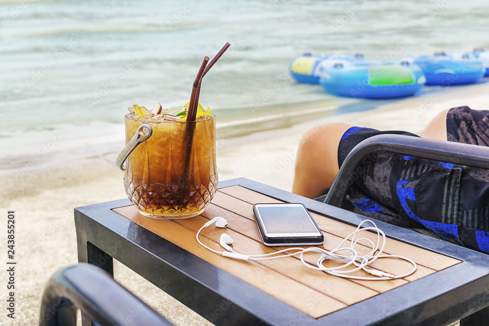 Fototapeta premium Man sitting at the beach with cocktail bucket, cell phone and earpods on table. Blurred sea and inflatable swimming rings in background. Vacation, travel concept