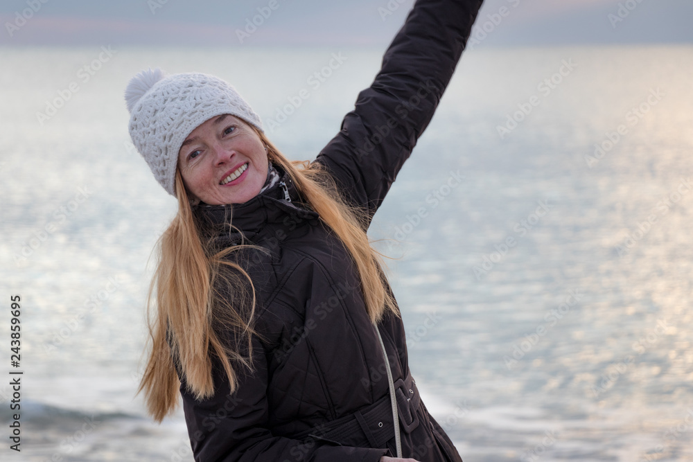 Smiling mature woman, with beautiful long hair, wearing a white knitted cap, at the beach enjoys a healthy active lifestyle in winter time. Winter mood. Copy space. Smiling mature woman, with beautiful long hair, wearing a white knitted cap, at the beach enjoys a healthy active lifestyle in winter time. Winter mood. Copy space.