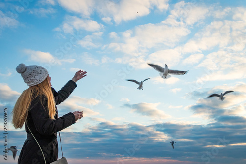 Happy mature woman, with beautiful long hair, wearing a white knitted cap, feeds the seagulls on the beach against the blue sky. Autumn mood. Copy space. © Alexander