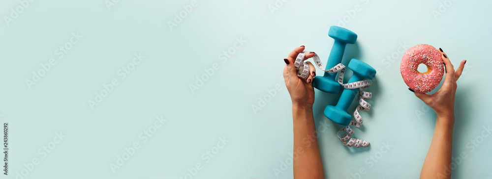 Female hand holding sweet donut, measuring tape, dumbbells over blue background. Top view, flat lay. Weight lost, sport, fitness, diet concept. Banner with copy space. - obrazy, fototapety, plakaty 
