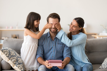 Loving wife and kid daughter making surprise to smiling dad receiving gift on fathers day, family closing eyes of excited daddy preparing present congratulating celebrating happy birthday at home