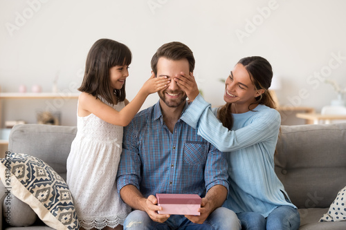 Loving wife and kid daughter making surprise to smiling dad receiving gift on fathers day, family closing eyes of excited daddy preparing present congratulating celebrating happy birthday at home