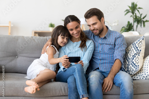 Smiling mother holding smartphone taking family selfie, watching video, making online call looking at phone screen, shopping in mobile app, recording vlog embracing child daughter and husband at home
