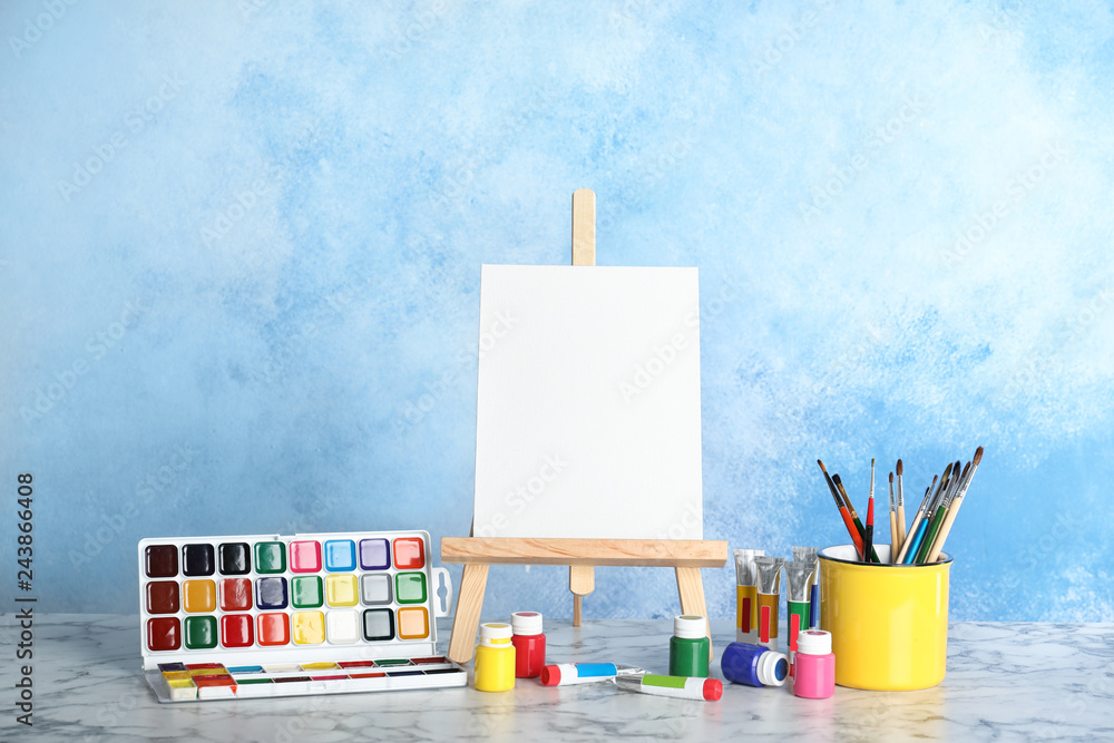 Obraz premium Wooden easel with blank canvas board and painting tools for children on table near color wall