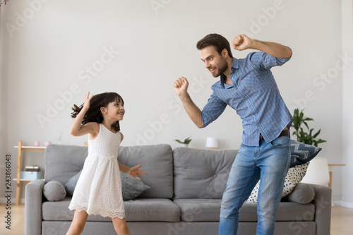 Happy dad and little funny daughter laughing dancing in living room, cheerful daddy and cute child girl having fun together, active kid enjoying copying fathers moves playing with parent at home photo