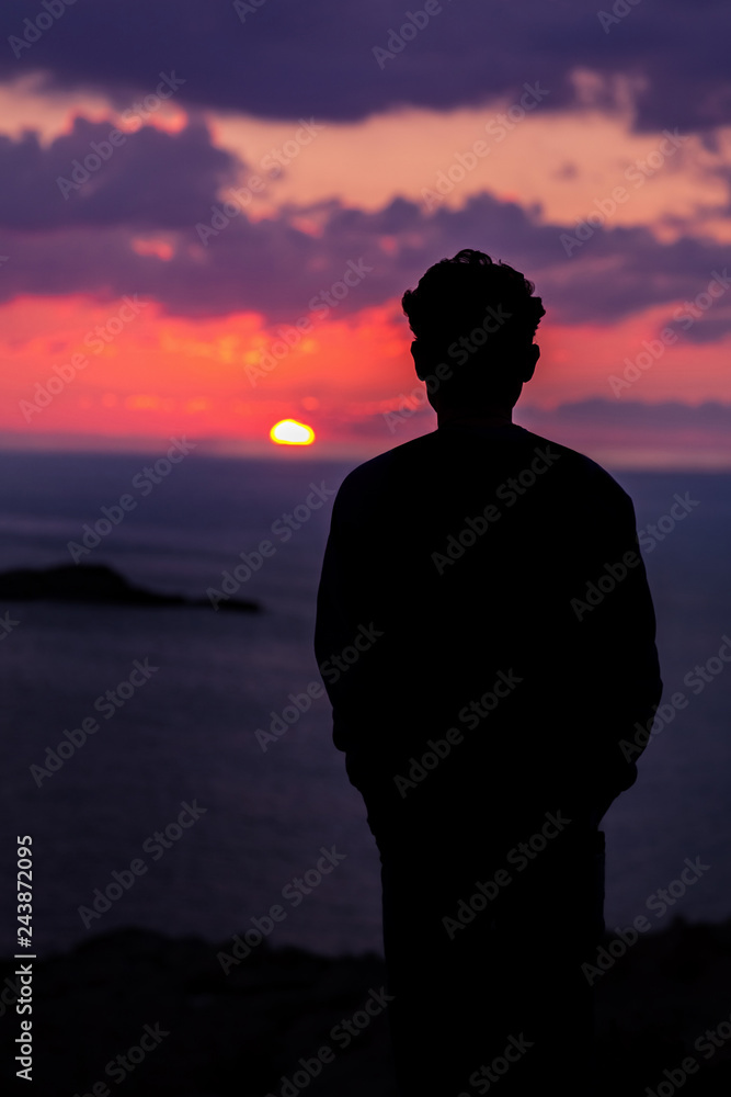 Silhouette of a young man standing on the edge of the cliff and looking at the sun that is setting over the sea