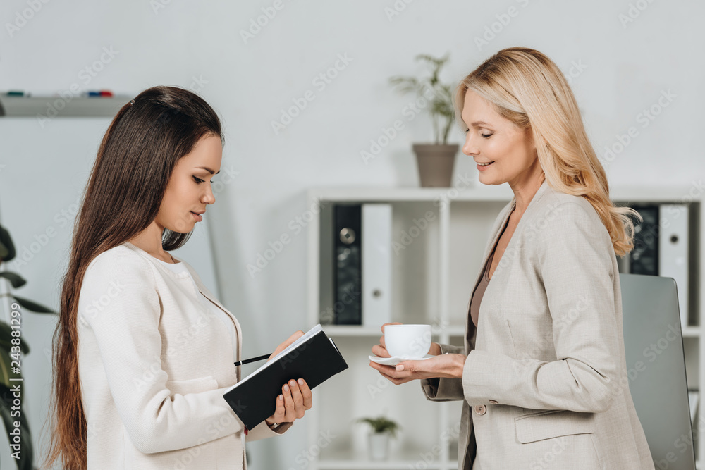 side view of smiling business mentor with cup of coffee looking at young female colleague taking notes in notebook