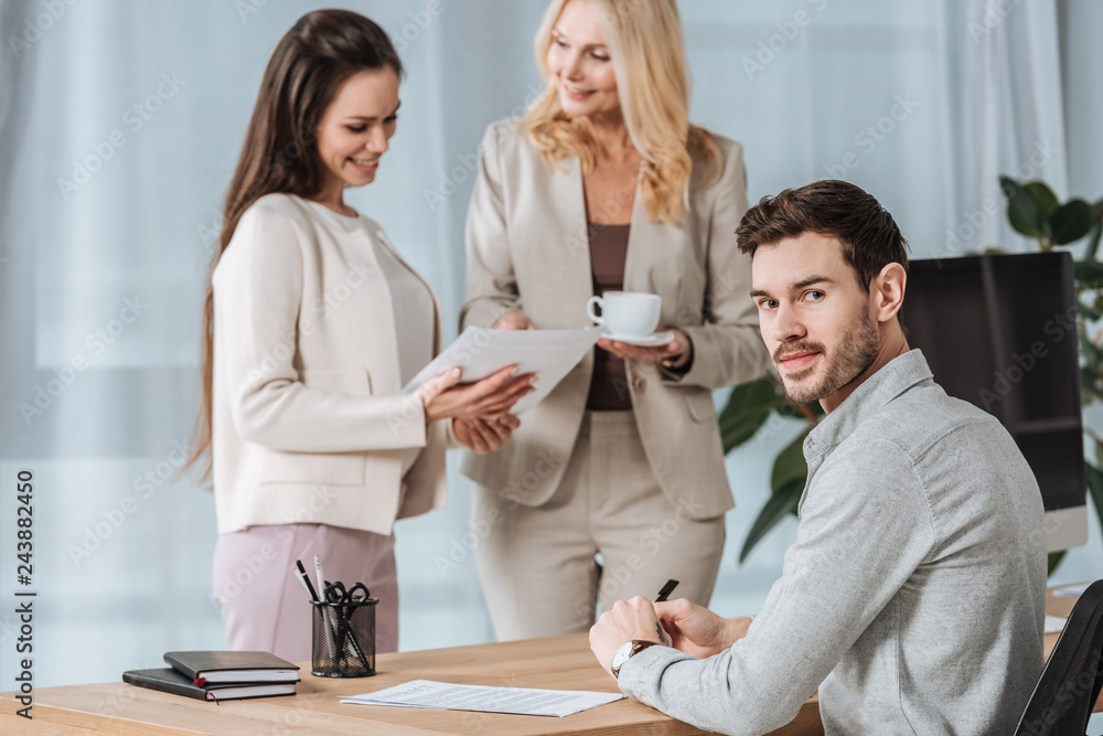 smiling businesswomen talking and young businessman looking at camera in office smiling businesswomen talking and young businessman looking at camera in office