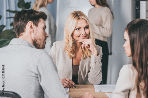 Fotografia beautiful smiling business mentor sitting with hand on chin and looking at young
