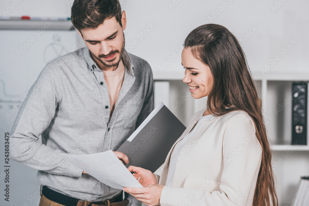 Fototapeta premium young business mentor holding folder and discussing papers with female colleague in office