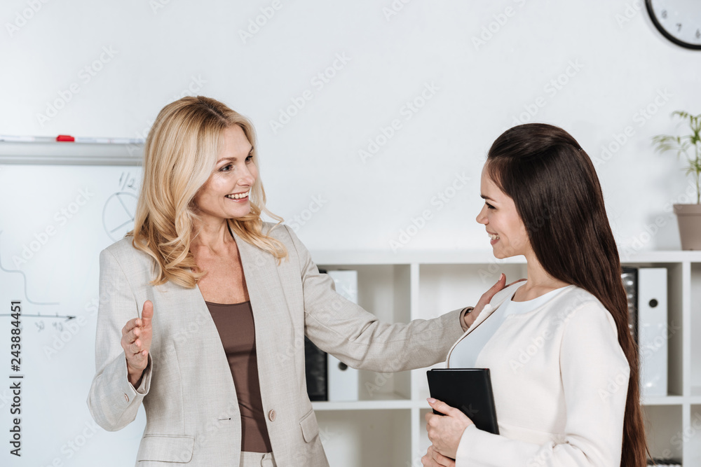 young businesswoman with notebook and cheerful mature mentor smiling each other in office young businesswoman with notebook and cheerful mature mentor smiling each other in office