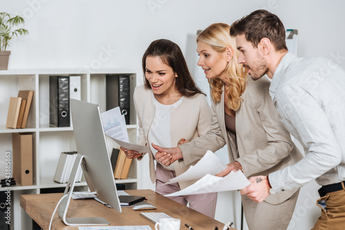 smiling business mentor with young colleagues holding papers and using desktop computer in office