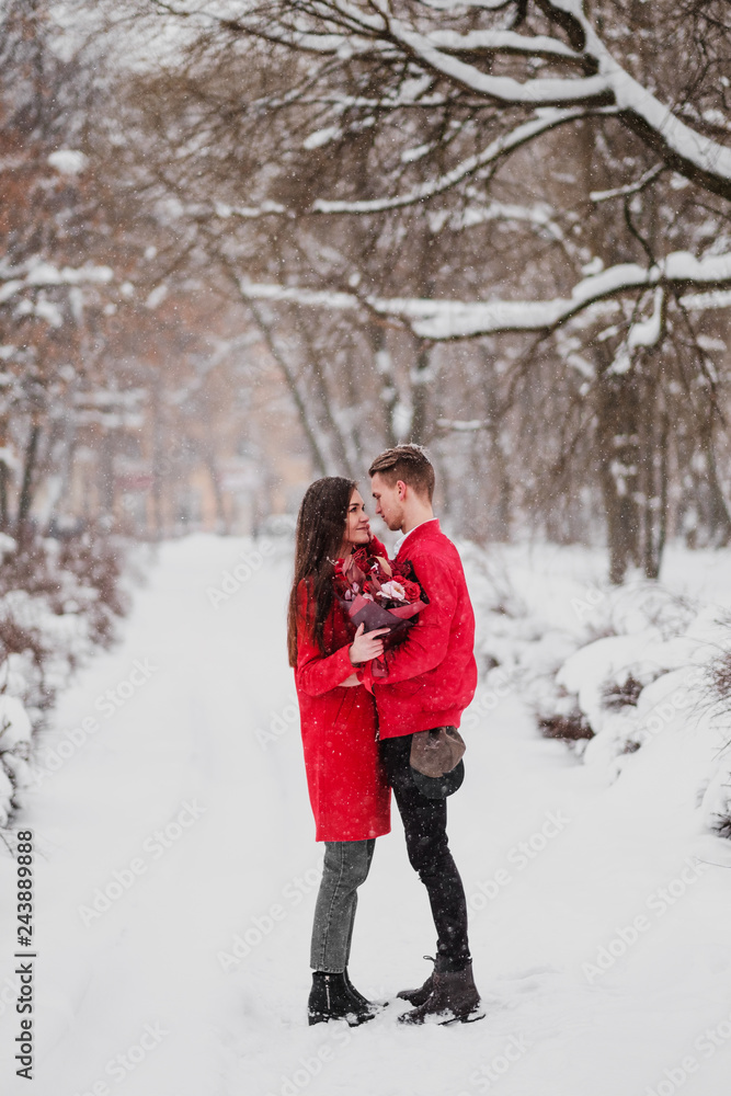 Fototapeta premium A date of lovers with my park in the winter. A bouquet of red flowers, walk, hug, kiss, laugh in a romantic setting. Portrait of a couple, husband and wife. Red jackets and coats.
