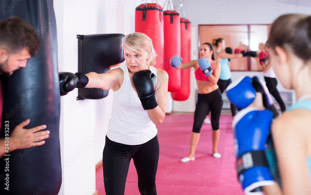 active mature female is doing exercises with punching bag in gym