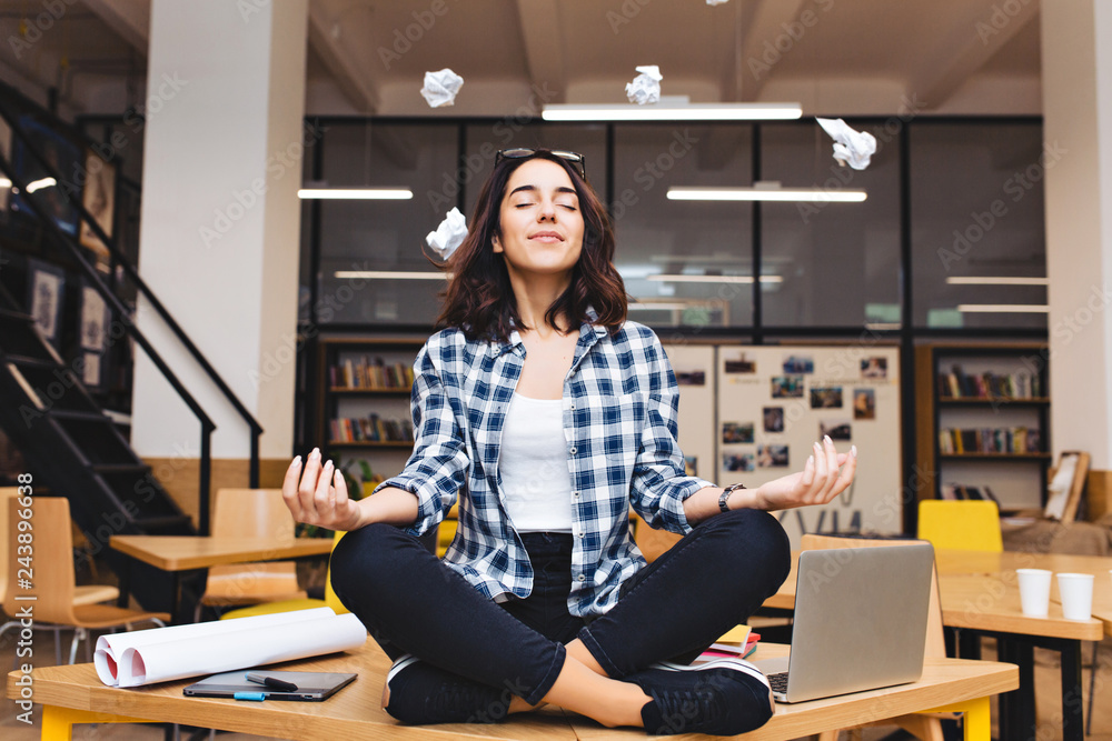 Young joyful brunette woman having meditation on table surround work stuff and flying papers. Cheerful mood, taking a break, at  work, studying, relaxation, smiling with closed eyes - obrazy, fototapety, plakaty 