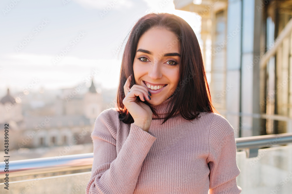 Fototapeta premium Portrait joyful smiled brunette young woman in sunny morning chilling on terrace. Smiling to camera, expressing positive face emotions, success, enjoying