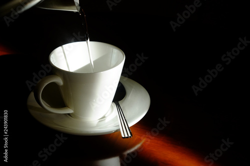 Filling hot water from kettle to empty white glass with saucer and metal spoon in the dark corner with beautiful sunlight on wooden glossy table