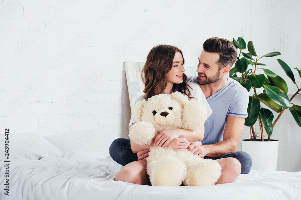 happy couple embracing teddy bear while sitting on bed with white bedding happy couple embracing teddy bear while sitting on bed with white bedding