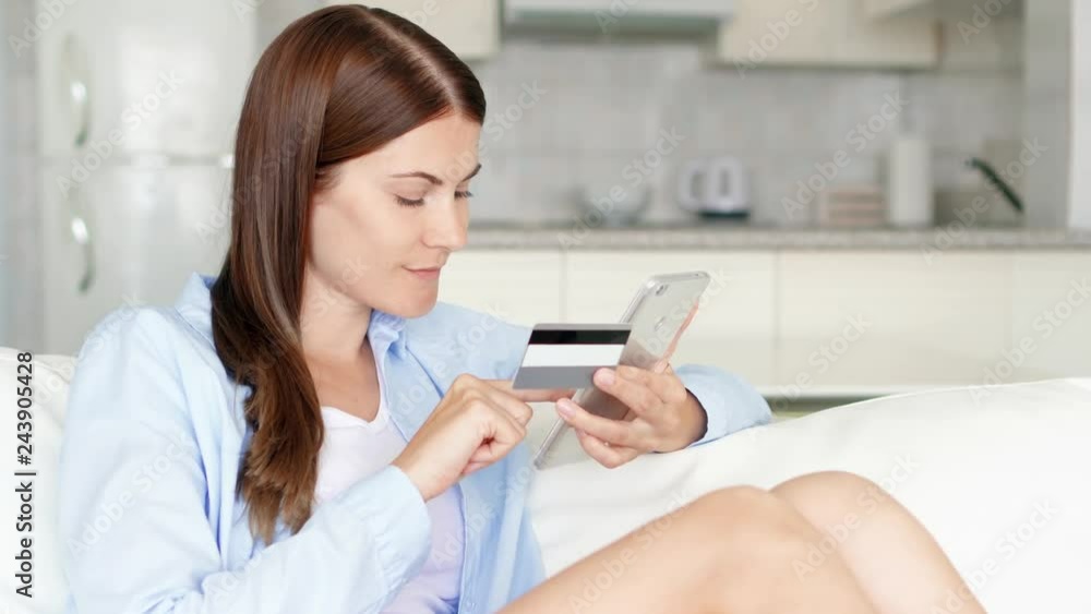 Young brunette woman with brown eyes sitting on sofa in living room using smartphone. Female housewife shopping online with credit card on mobile phone. Consumerism and internet online purchases Young brunette woman with brown eyes sitting on sofa in living room using smartphone. Female housewife shopping online with credit card on mobile phone. Consumerism and internet online purchases