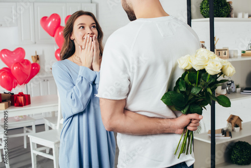 partial view of a young man holding bouquet of roses behind back while smiling girlfriend waiting for surprise