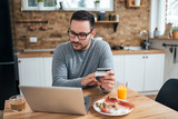 Handsome man at the kitchen table with breakfast holding credit card and looking at laptop.