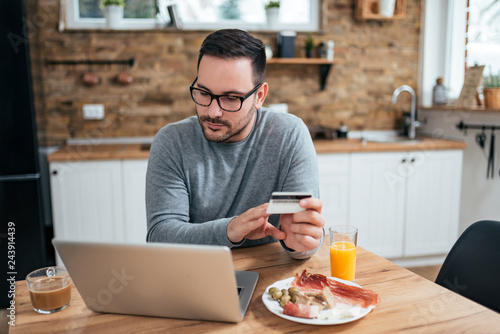 Handsome man at the kitchen table with breakfast holding credit card and looking at laptop.