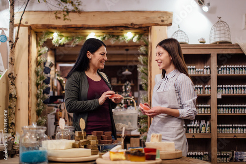 Cheerful shopkeeper helping customer in packaging free shop. Zero waste shopping - woman buying natural homemade soap at package free store.