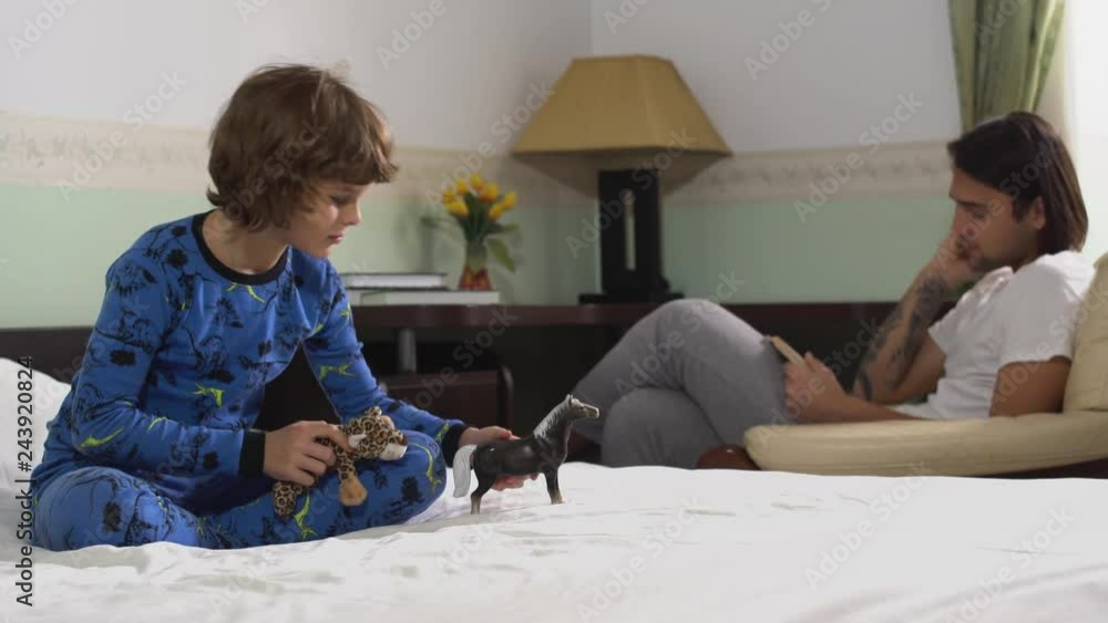 One younger kid playing on the bed with horse and tiger toys and older brother reading a book in arm chair. Brothers relationship. One younger kid playing on the bed with horse and tiger toys and older brother reading a book in arm chair. Brothers relationship.