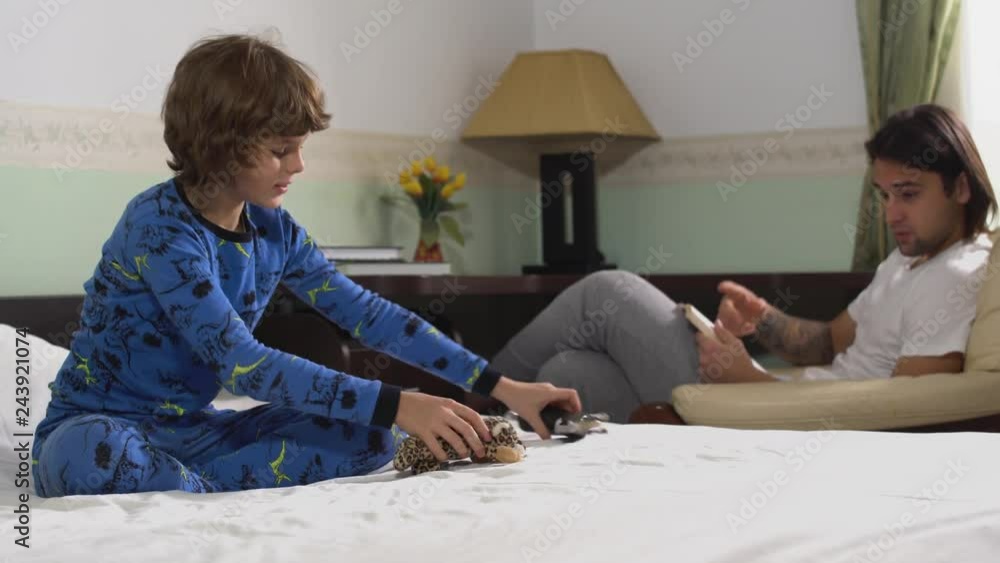 Older brother reading a book sitting in the chair asks the younger brad to play with toys while sitting on the bed to keep himself quiet. Brothers relationship. Older brother reading a book sitting in the chair asks the younger brad to play with toys while sitting on the bed to keep himself quiet. Brothers relationship.