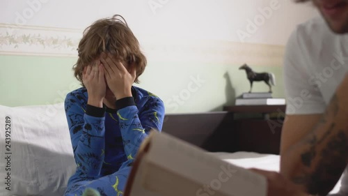 Kid sitting on big bed and rub his eyes because boy very tired from book which his brother read for him. Brothers relationship. photo