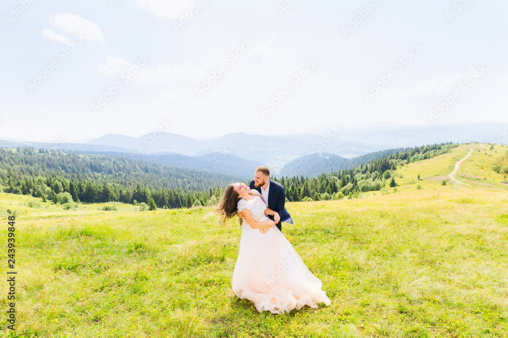 Newlyweds smile and hug each other among the meadow on top of the mountain against the background of panorama of the mountains.
