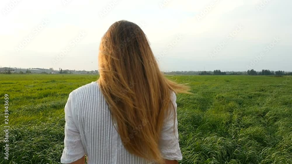 Beautiful woman in white dress walks on the green field. Wind blowing hair. Grassy summerfield on sunset Beautiful woman in white dress walks on the green field. Wind blowing hair. Grassy summerfield on sunset